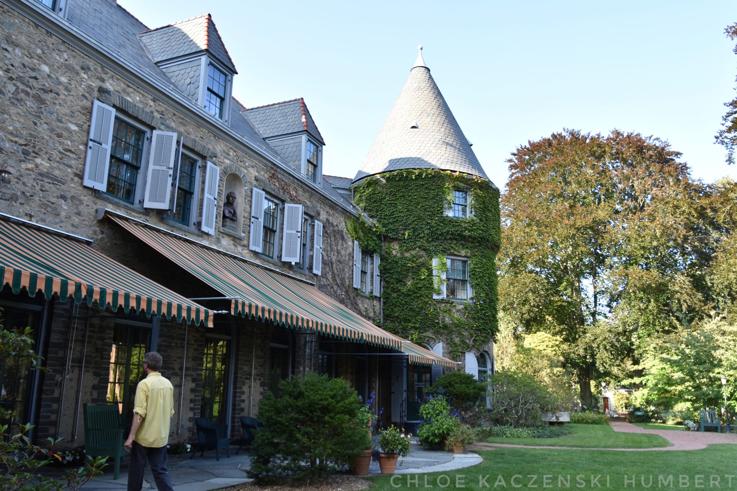 Grey Towers National Historic Site aka Gifford Pinchot House in Dingman Township Milford Pennsylvania USA Photo by Chloe Kaczenski Humbert. The photo shows a person walking on the grounds of a castle like stone mansion with a cylindrical turret covered in vines with pointed roof, and an awning over a stone patio shades large windowed doors, the yard has trees, bushes, and a manicured lawn.